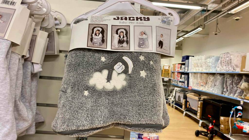 In einem Geschäft ist eine Einschlafdecke im Test, eine graue Jacky Plüschdecke inklusive Fußsack auf einem Bügel hängend, an einem Verkaufsregal ausgestellt.