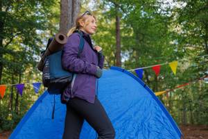 Jade-Yogamatte-Test: Eine Frau in Wanderkleidung geht vor einem Zelt und hat eine Yogamatte auf ihrem Rucksack befestigt. 