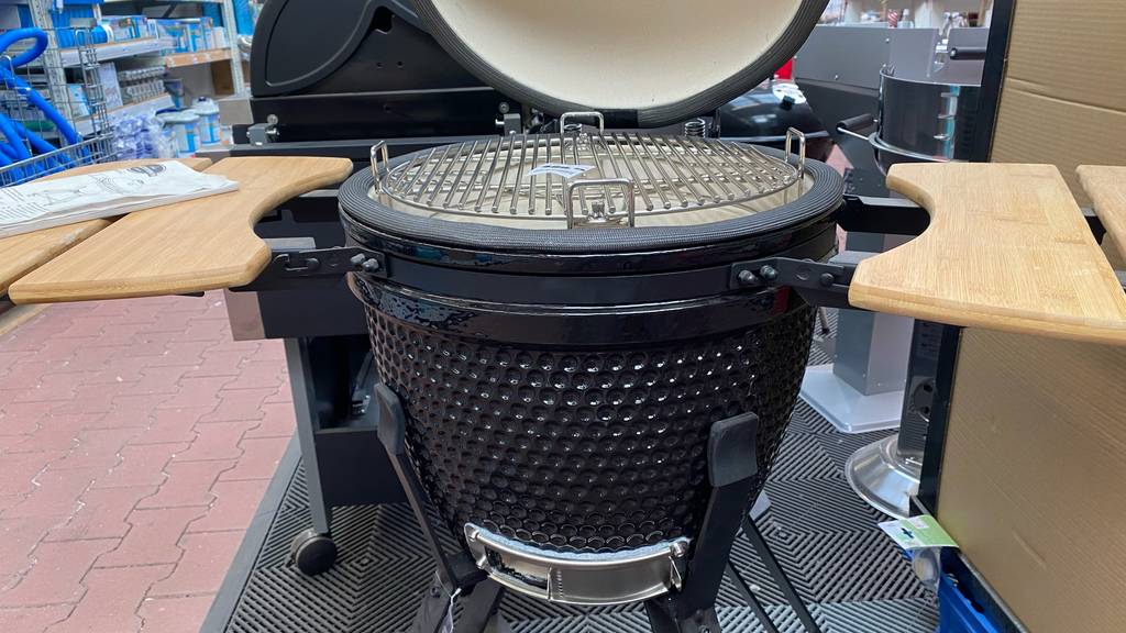 Keramikgrill im Test: ein schwarzes, rundes Modell mit seitlichen Holz-Ablageflächen steht geöffnet in einem Verkaufsbereich.