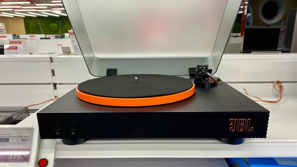 Ein getesteter Bluetooth-Plattenspieler von vorne mit geöffneter Klappe und Filz auf dem Plattenteller und Logo auf der rechten Seite, in einem Verkaufregal mit Preisangabe.