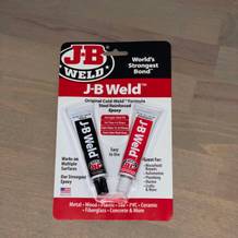Die Verpackung des Produkts J-B Weld ist frontal auf einem Holztisch zu sehen. Sie zeigt die beiden Klebstoff-Komponenten in Tubenform mit Produktinformationen.
