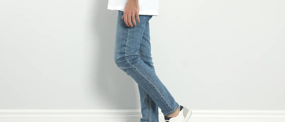 Jeans für Herren im Test: Mann mit einer blauen Jeans, einem weißen T-Shirt und weißen Schuhen steht vor einer weißen Wand.