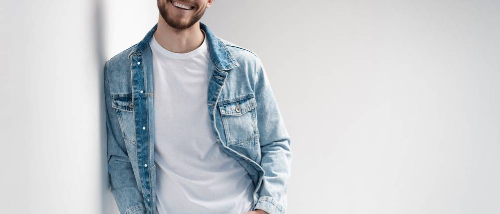 Jeansjacken für Herren im Test: Mann mit weißem T-Shirt und hellblauer Jeansjacke lehnt sich an einer weißen Wand an.