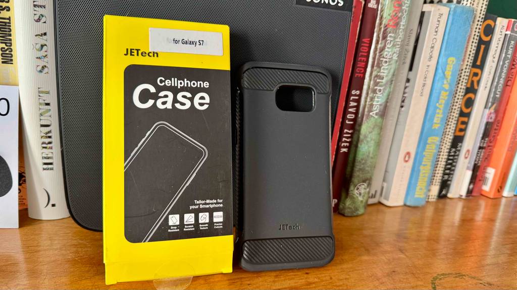 Getestet: Die Samsung-Galaxy-S7-Hülle in Schwarz lehnt neben ihrer schwarz-gelben Verpackung an einer Bücherwand auf einem Holzregal.