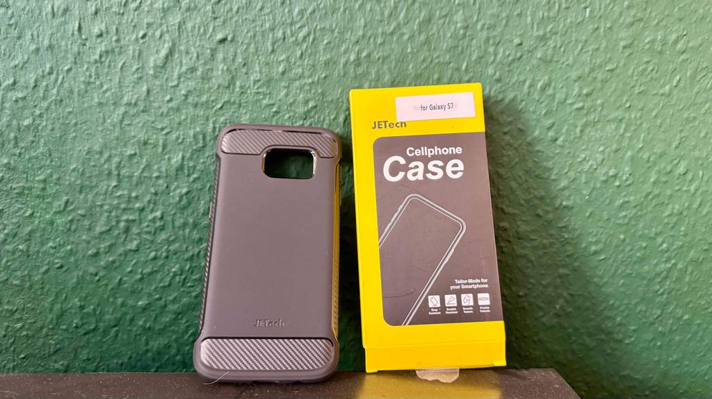 Im Test: Die Samsung-Galaxy-S7-Hülle in Schwarz lehnt neben ihrer schwarz-gelben Verpackung an einer grünen Wand.