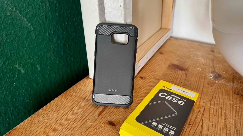 Die schwarze Samsung-Galaxy-S7-Hülle im Test lehnt neben ihrer auf einer Holzoberfläche liegenden schwarz-gelben Verpackung an zwei weißen Leinwänden.