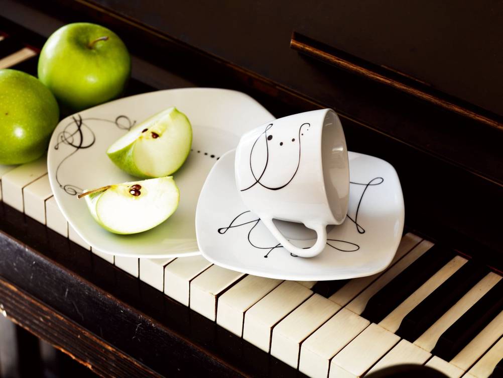 Dekor auf dem Kombiservice getestet. Kuchenteller, Untertasse und Tasse eines weißen Services mit schwarzen Dekorlinien ist gemeinsam mit grünen Äpfeln auf einer Klaviertastatur abgestellt.