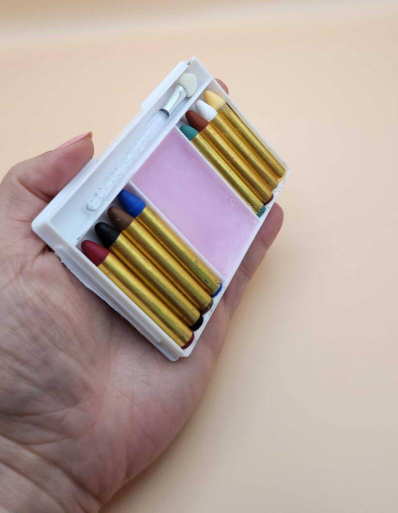 Kinderschminke-Test: Eine Hand hält eine Packung, in der sich mehrere farbige Stifte befinden, vor einem beigefarbenen Hintergrund.