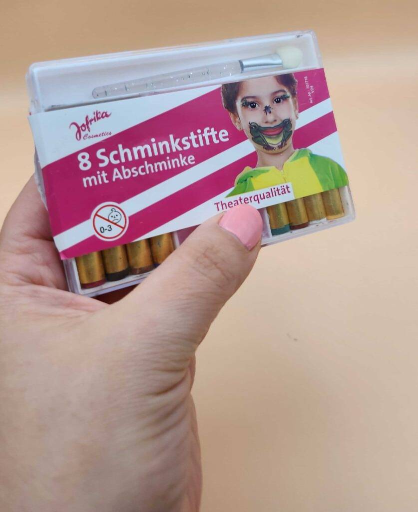 Kinderschminke im Test: Eine Hand hält eine Packung vor einer beigefarbenen Oberfläche.