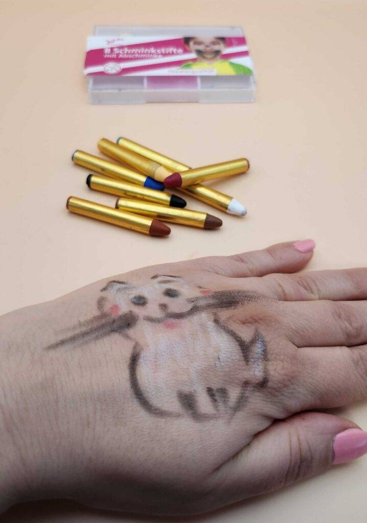 Kinderschminke-Test: Auf einem Handrücken ist eine gemalte Katze zu sehen. Schminkstifte liegen im Hintergrund.