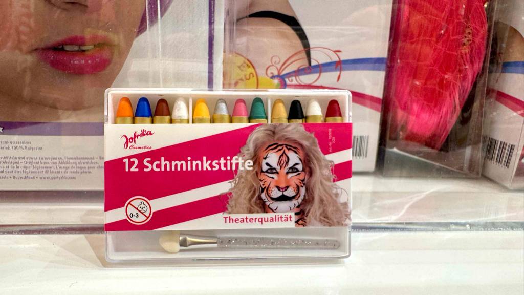 Im Test: Eine Packung mit 12 Schminkstiften steht vor verpackten Perrücken.