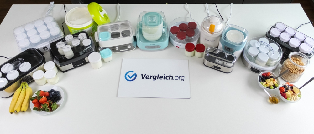 Zahlreiche Joghurtbereiter stehen hinter einem Schild mit dem Schriftzug vergleich.org