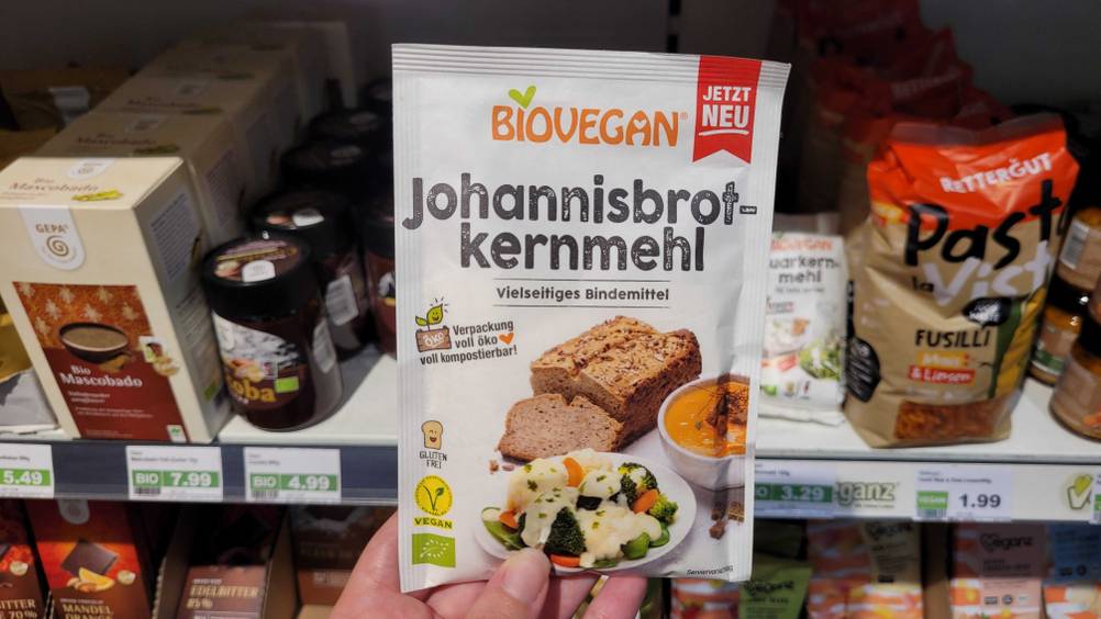 Eine Hand hält eine Packung des getesteten Johannisbrotkernmehls vor ein Regal mit anderen Produkten.