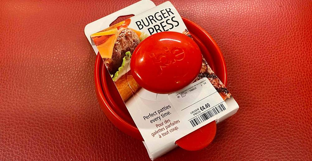 Burgerpressen-Test: Nahaufnahme einer roten Hamburgerpresse inklusive Verkaufskarton und Preisschild auf einer roten Oberfläche liegend.