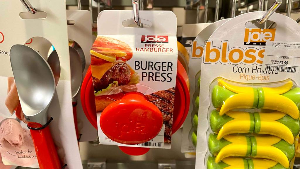 Eine rote Joie Burgerpresse im Test hängt an seinem weißen Verpackungskarton an einem Verkaufsstand neben weiterem Haushaltszubehör.