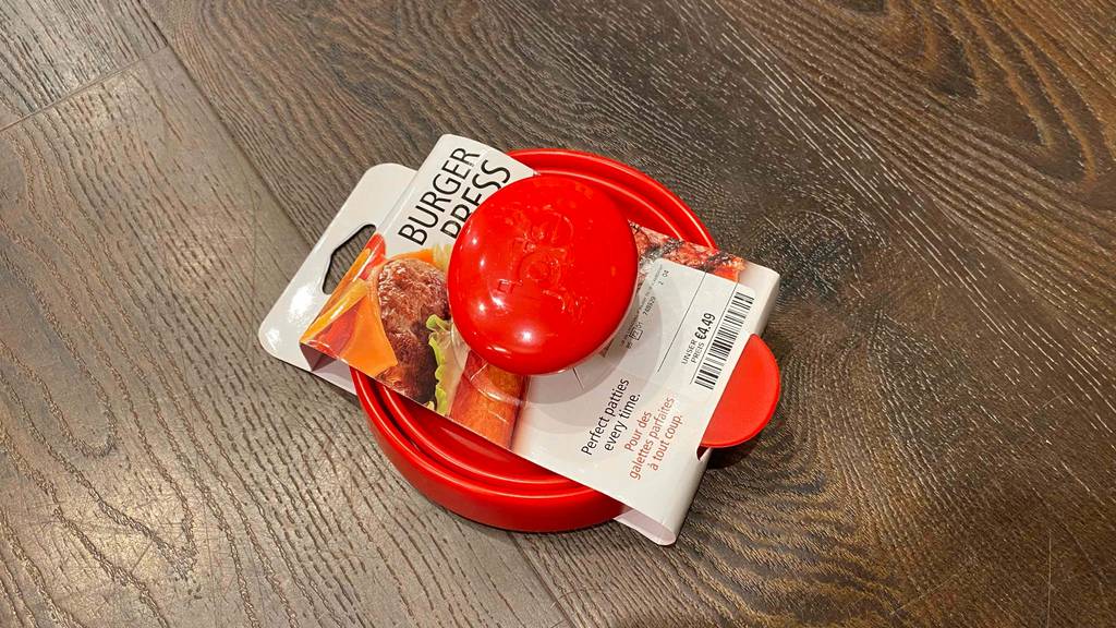 Eine rote Burgerpresse im Test liegt im weißen Verpackungskarton inklusive Preisschild auf einer dunklen Holzfläche.