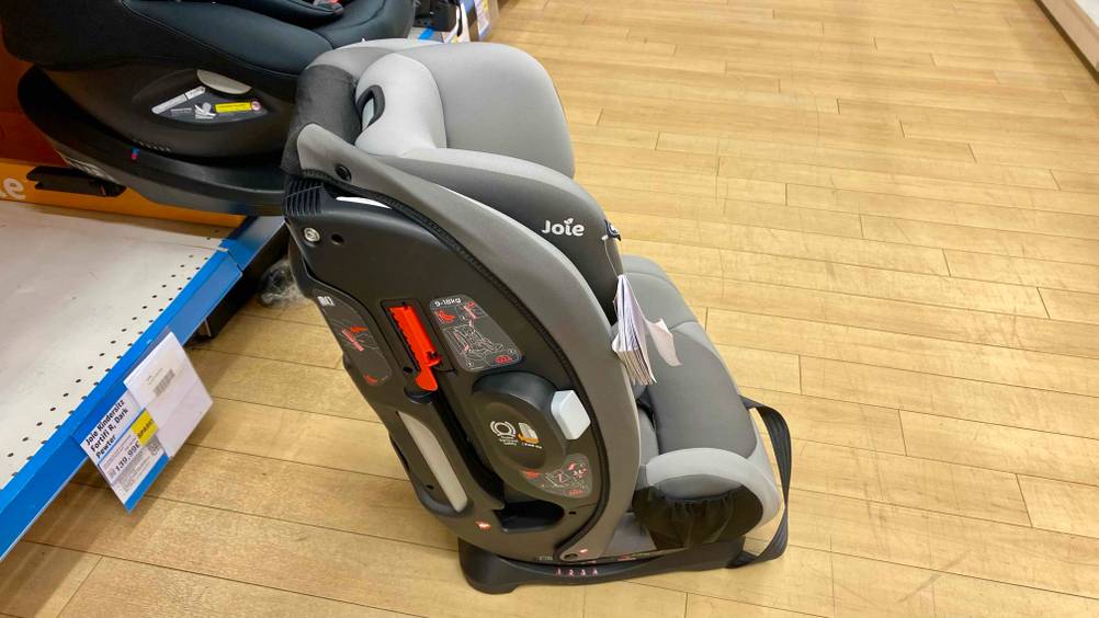 Ein getesteter Kindersitz ab 1 Jahr von der Seite mit Infografiken zur Befestigung auf einem Holzboden vor einem Verkaufsregal.