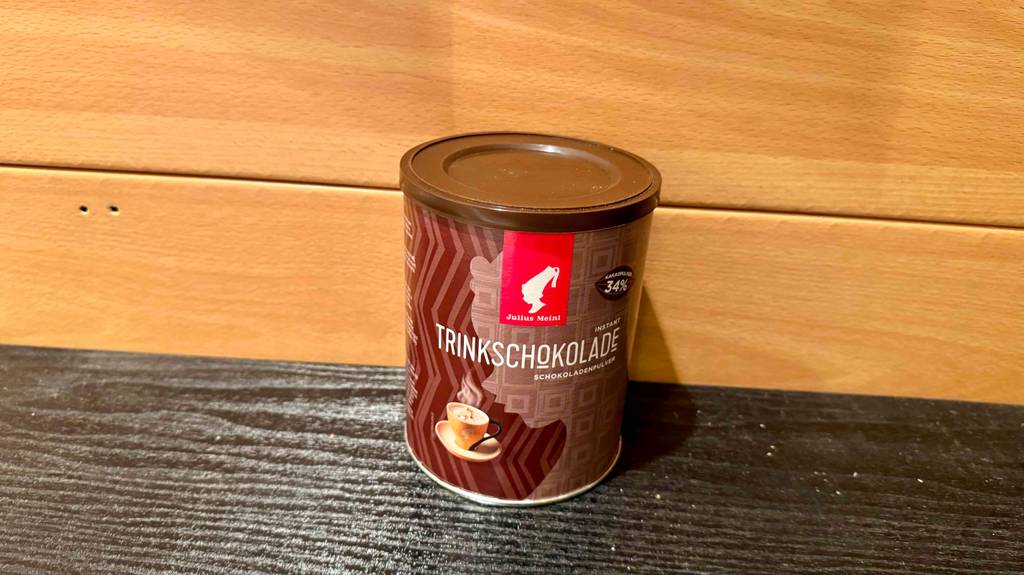 Eine wiederverschließbare Dose mit Pulver für Trinkschokolade im Test steht vor einer Holzwand.