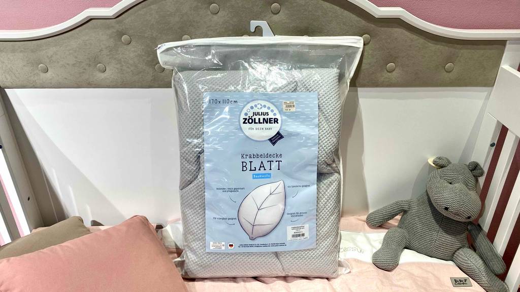 Hellgraue getestete Krabbeldecke von Julius Zöllner befindet sich in der Originalverpackung auf einem Kinderbett neben einem Kuscheltier.