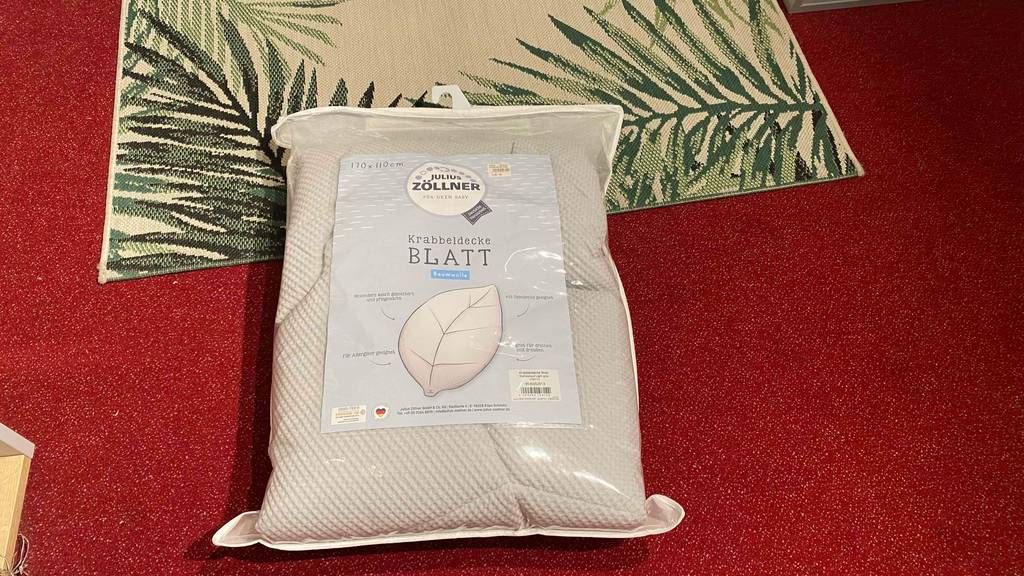 Beigefarbige Julius Zöllner Krabbeldecke im Test liegt in der Originalverpackung auf einem roten Teppich.