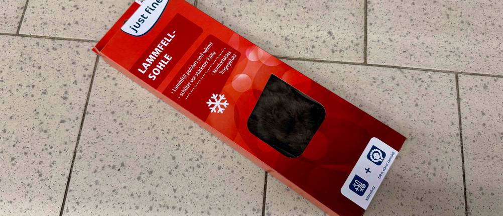 Eine getestete Lammfellsohle liegt in der roten Verpackung auf einem Fliesenboden.