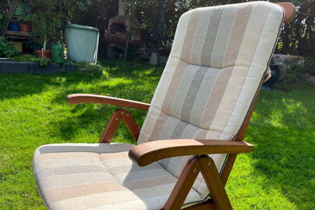 Eine Holz-Gartenstuhl mit einer hellen Auflage steht in einem Garten.