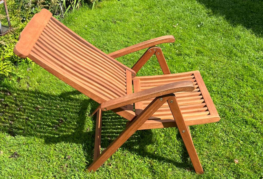 Ein getesteter Holz-Gartenstuhl mit herabgelassener, verstellten Rückenlehne von der Seite betrachtet, auf einem Rasen.
