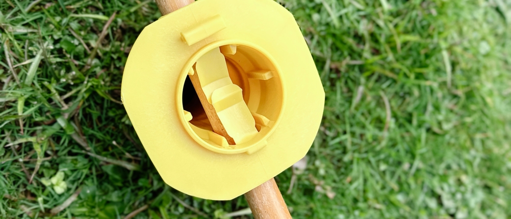 Gelbe Kunststoffhalterung der Juwel Novaplus 600 Lift liegt lose auf einem Holzstab auf einer Rasenfläche.