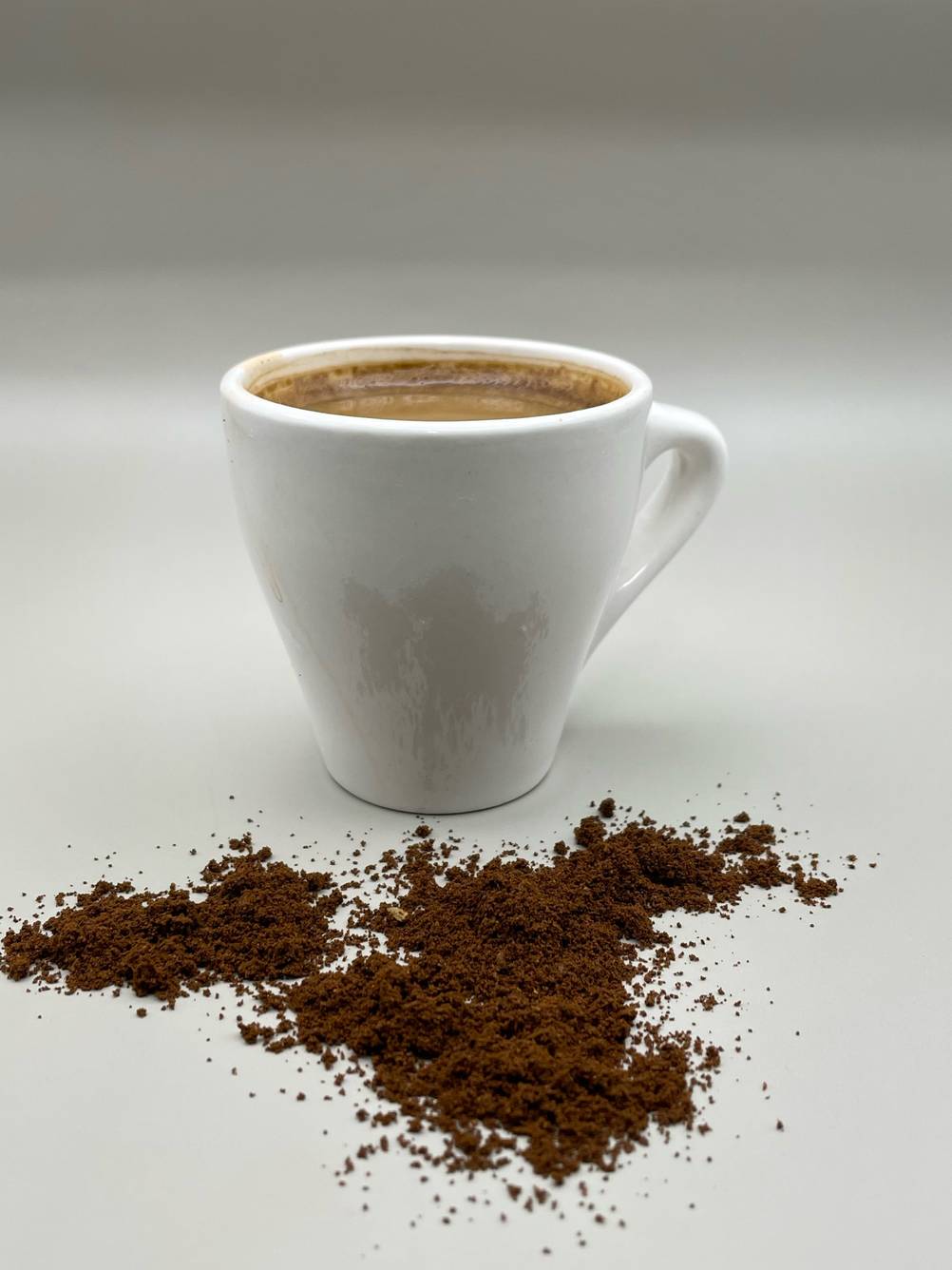 Vor einer Tasse des Kaffees in Trommelröstung liegt der Kaffe auf einer hellen Fläche.