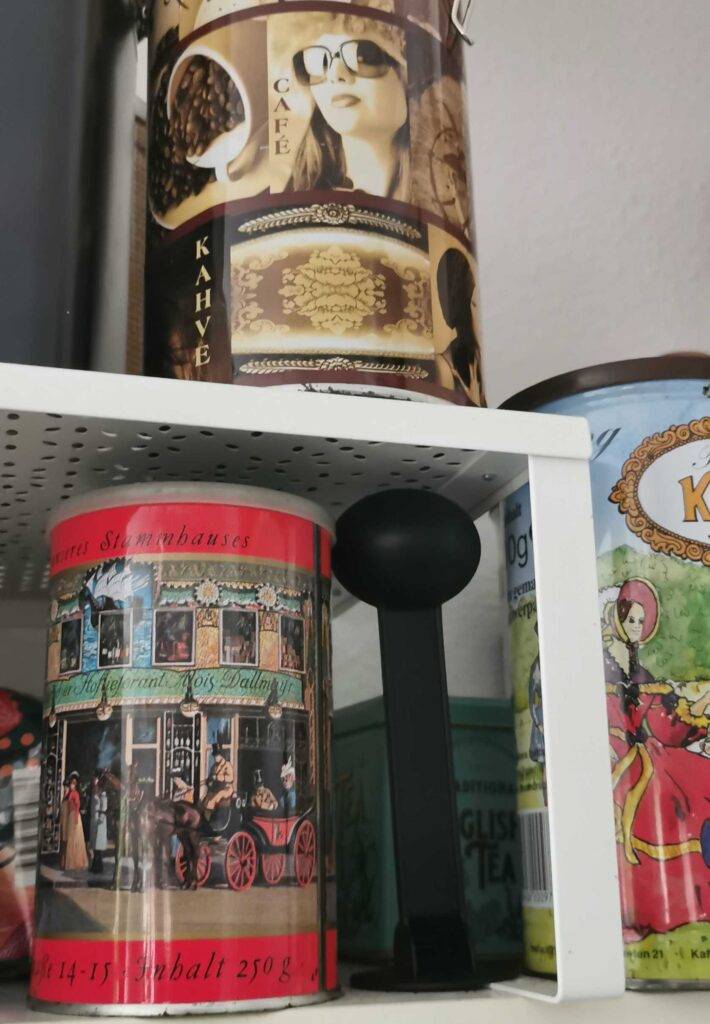 Mehrere bunte getestete Kaffeedosen in unterschiedlichem Design stehen neben einem Kaffeelöffel auf einem Regal.