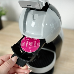 Eine Hand hält eine Dolce Gusto Espresso-Kapsel unterhalb der geöffneten Krups Mini Me. Die Kapsel befindet sich im Auszug.