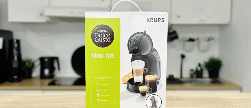 Die Originalverpackung der Krups Nescafé Dolce Gusto Mini Me KP120H steht auf einem Tisch. Das Modell ist mit Getränkebeispielen dargestellt.