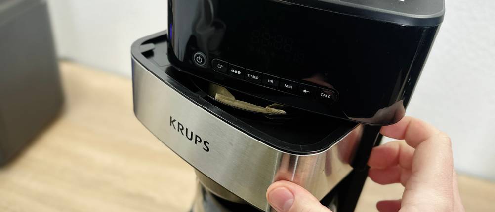 Die Vorderseite der Krups-KM8328-Kaffeemaschine wird mit einer Hand geöffnet. Das Bedienfeld und der Filterhalter sind dabei deutlich zu erkennen.