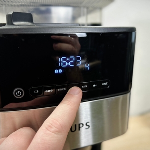 Ein Finger tippt auf das Display der Krups KM8328, um die Minutenanzahl einzustellen. Die Uhrzeit und Symbole sind deutlich lesbar.