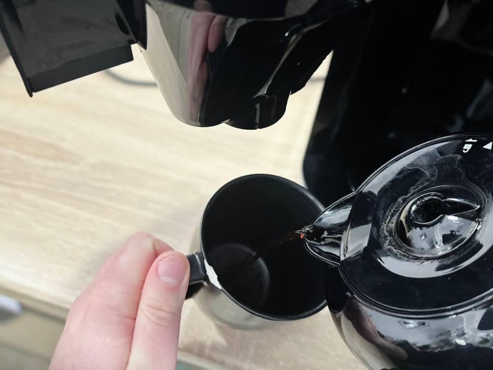 Kaffee wird aus der Glaskanne der Krups KM8328 in eine schwarze Tasse gegossen. Die Hand hält die Kanne sicher am Griff.