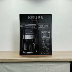 Die Vorderseite der Verpackung der Krups KM8328 ist komplett zu sehen. Abgebildet sind Produktname, Kaffeemaschine, Funktionen und Kaffeebohnen als Dekoelemente.