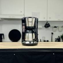 Die Russell Hobbs Adventure Thermo-Kaffeemaschine steht mittig auf einer Holzplatte. Die Küche ist im Hintergrund sichtbar.