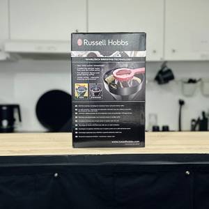 Die Rückseite der Verpackung der Russell Hobbs Adventure Kaffeemaschine mit technischen Infos und Feature-Icons steht auf einer Holzfläche.