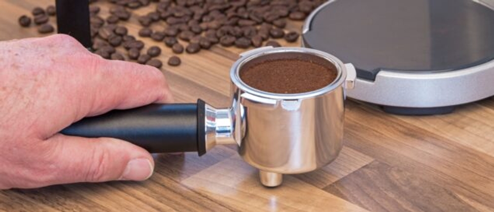 Eine Hand hält einen gefüllten Siebträger vor Kaffeebohnen und einer Kaffeemaschine.