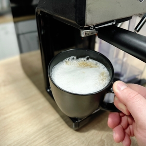 Eine Tasse mit frisch gebrühtem Kaffee und Milchschaum steht unter dem Auslauf der Breville Prima Latte II VCF108X. Eine Hand hält die Tasse am Griff.
