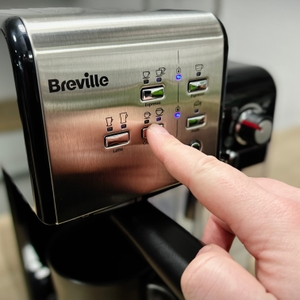 Ein Finger tippt auf das Bedienfeld der Breville Prima Latte II VCF108X. Die Taste für Cappuccino ist aktiviert, daneben sind weitere Getränkewahlen sichtbar.