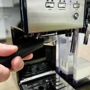 Eine Hand setzt den Siebträger in die Breville Prima Latte II VCF108X ein. Im Hintergrund sind Milchbehälter und Tassenablage zu sehen.