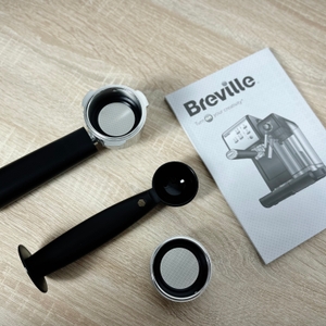 Das Zubehör der Breville Prima Latte II VCF108X liegt auf einem Tisch: Siebträger, Löffel mit Tamper und zwei Filtereinsätze, daneben die Anleitung.