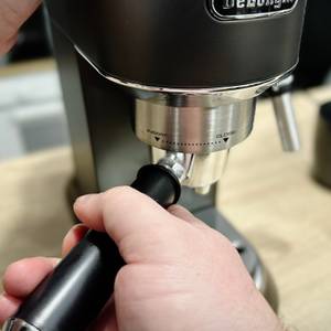 Eine Hand setzt den Siebträger in die Brühgruppe der De'Longhi Dedica Style EC 685.B ein. Die Maschine steht auf einer Holzoberfläche.