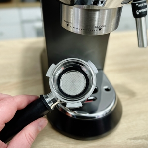 Ein leerer Siebträger wird vor die Brühgruppe der De'Longhi Dedica Style EC 685.B gehalten. Der Metallfiltereinsatz ist gut sichtbar.