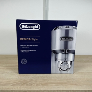 Die Originalverpackung der De'Longhi Dedica Style EC 685.B steht geschlossen auf einer Holzfläche. Das Design zeigt die Maschine frontal.