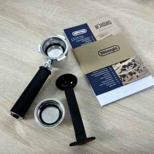 Der Lieferumfang der De'Longhi Dedica Style EC 685.B liegt auf einem Holztisch: Siebträger, Messlöffel mit Tamper, Siebe und Anleitung.