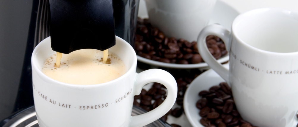 Kaffeepadmaschinen-Test: Kaffeepadmaschine bereitet einen Kaffee zu, daneben stehen noch zwei leere Tassen, die auf Untertassen stehen, umgeben von Kaffeebohnen.