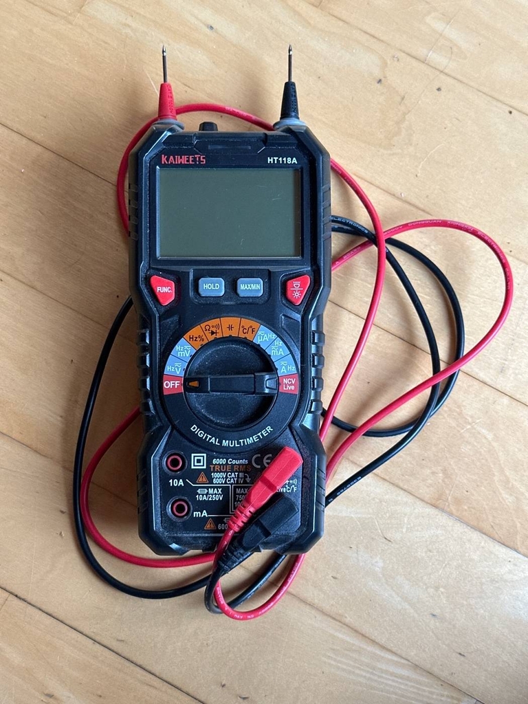 Multimeter im Test: Ansicht von oben auf ein ausgeschaltetes, digitales Kaiweets Messgerät in blauer Ausführung inklusive rotem sowie schwarzem Messkabel auf einem Holzboden.