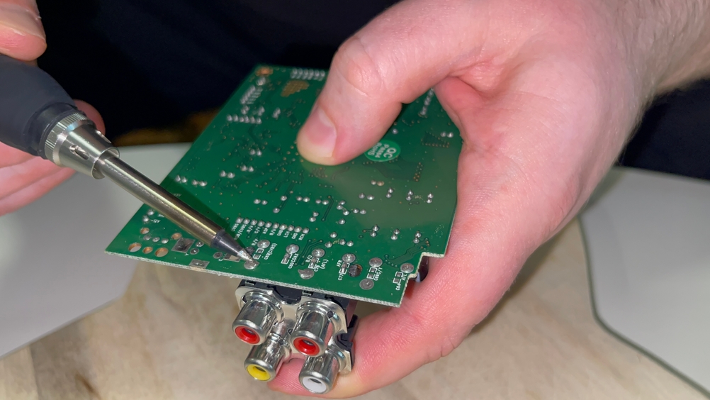 Ein Lötkolben berührt die Unterseite einer grünen Platine mit mehreren Lötstellen und Cinch-Anschlüssen. Die Platine wird dabei mit einer Hand gehalten, während die Lötspitze präzise angesetzt ist.